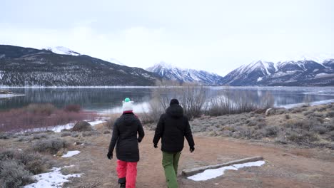 Couple-walking-towards-frozen-lake-with-snow-capped-mountains-in-the-background,-static