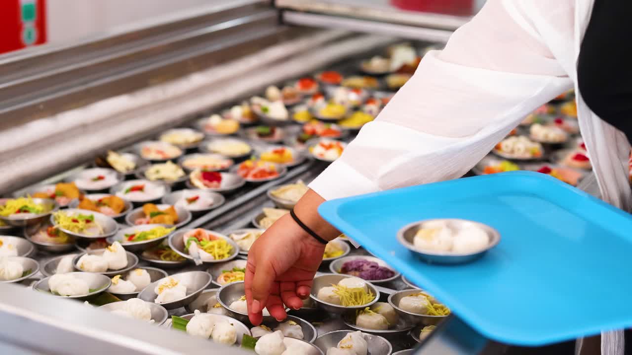 A person selects steamed dim sum from a vibrant display in a well-lit Phuket market, using a blue tray