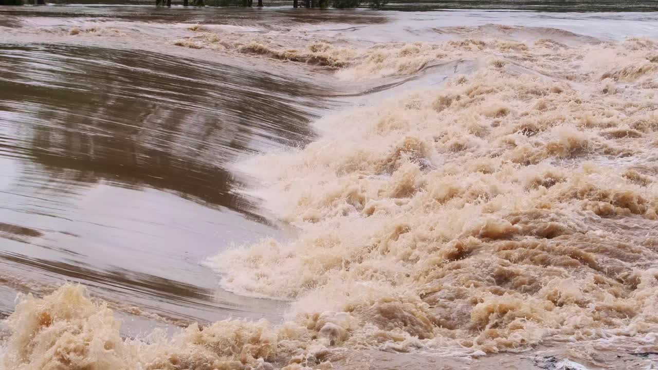 Close-up of brown floodwater flowing rapidly over the riverbanks on the Gold Coast in Queensland, Australia.