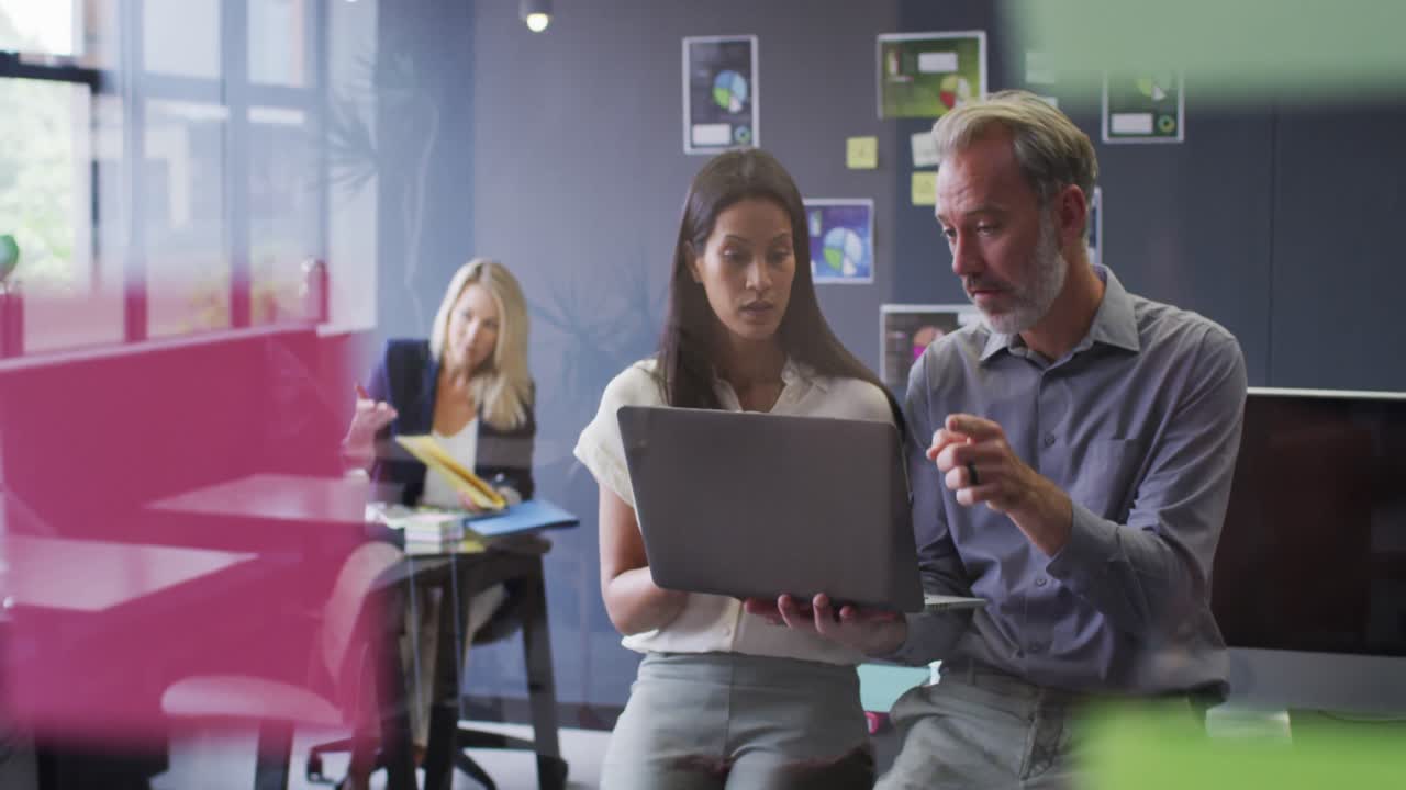 Two diverse business people using laptop talking in office and colleague working behind