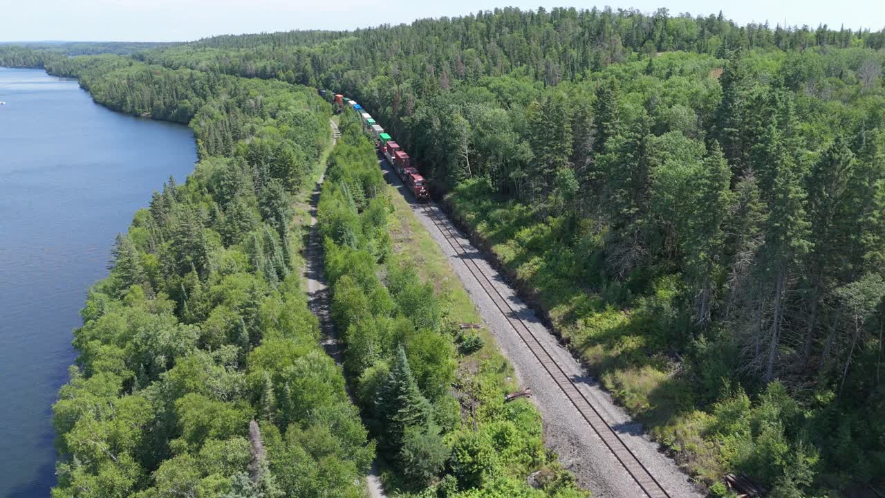 Aerial drone shot of a freight train traveling beside a blue lake through a lush green forest on a sunny summer day