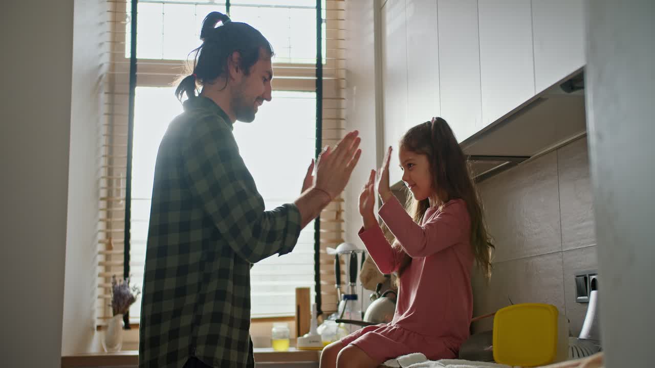 행복한 아버지, 체크 된 녹색 셔츠를 입은 갈색 남자가 분홍색 드레스를 입은 작은 갈색 딸과 함께 놀고, 현대적인 부에서 손을 박고 있습니다.