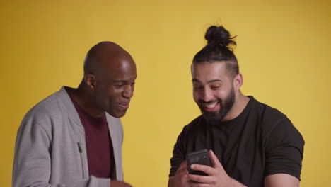 studio shot of two excited male friends with mobile phone celebrating winning money against yellow background 3