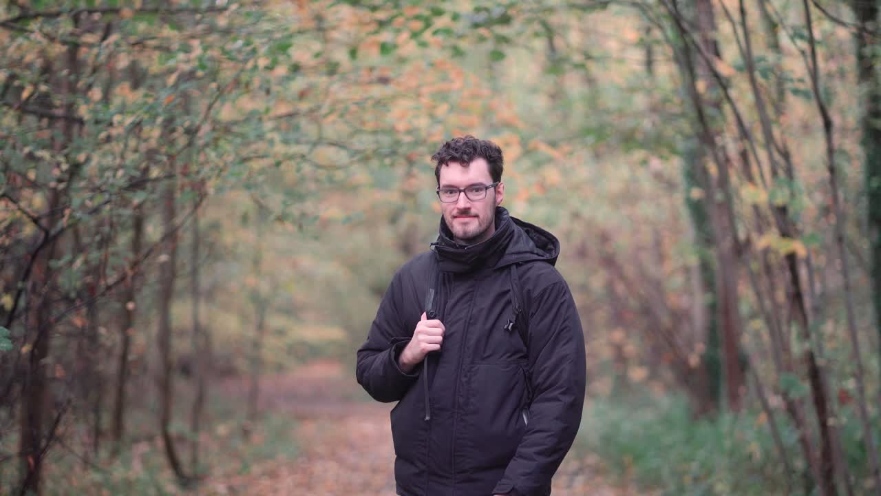 Engaging bearded European man with sharp glasses, standing in a mixed autumn forest, enthusiastically pointing and beckoning viewers closer, inviting them to join a nature exploration journey