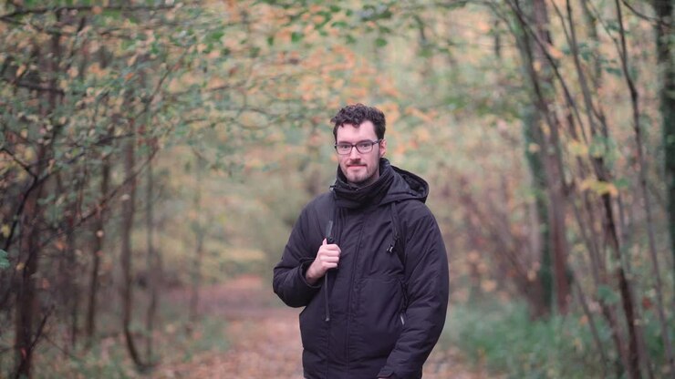Engaging bearded European man with sharp glasses, standing in a mixed autumn forest, enthusiastically pointing and beckoning viewers closer, inviting them to join a nature exploration journey