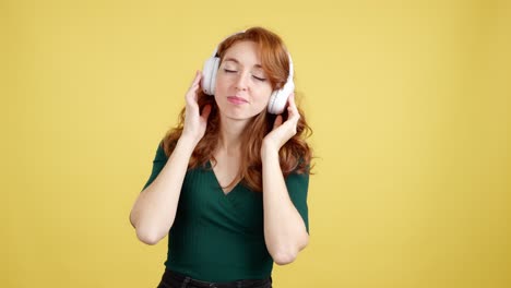 happy young woman with red hair dancing and enjoying music with headphones on a yellow background