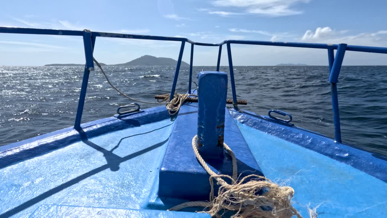 Forward-facing view from blue boat moving on sunlit ocean, approaching island, with gentle camera sway