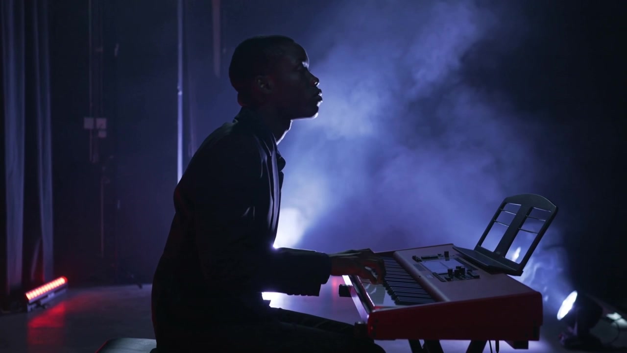 Playing keyboardist in dark clothes, sitting up, lifting head on cue at stage with red keyboard