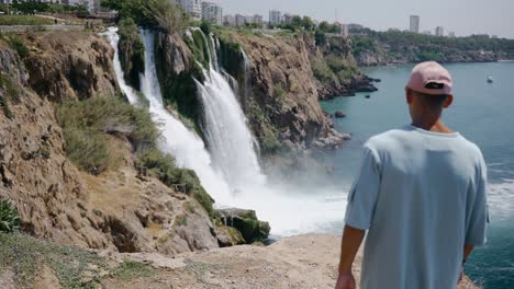 Latin-tourist-man-walking-towards-a-cliff-with-waterfalls-in-the-background,-touristic-hiking-landscape