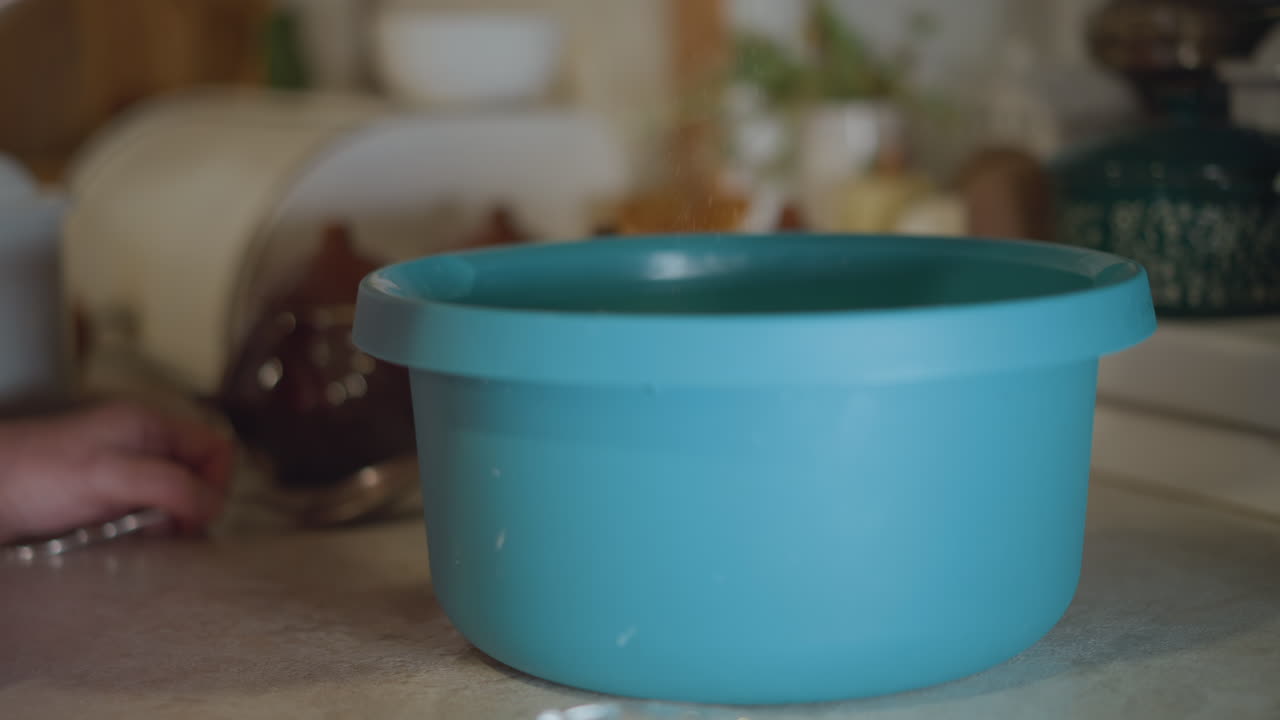 Pequeno prato de vidro perto de tigela azul. Mão coloca pequeno prato de vidro ao lado de tigela grande azul de mistura na bancada, luz suave da manhã e utensílios de cozinha vintage emoldurando a cena. Cozinheiro caseiro medindo uma pitada de sal