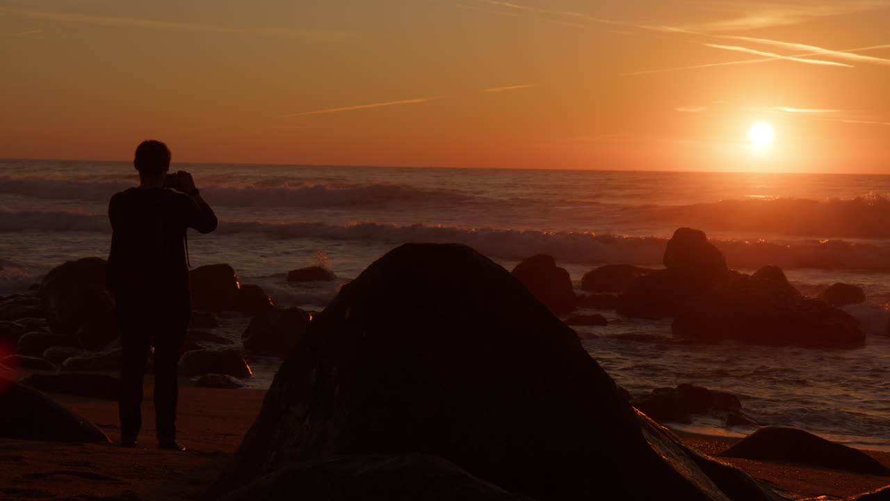 Siluet Seorang Pria di Pantai Berbatu Mengambil Gambar Matahari Terbenam Megah di Atas Cakrawala Samudra di Porto, Portugal. Bidikan Statis