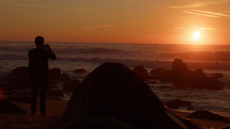silhouette of a man on rocky shore taking shots of majestic sunset above the horizon of ocean in porto, portugal. static shot