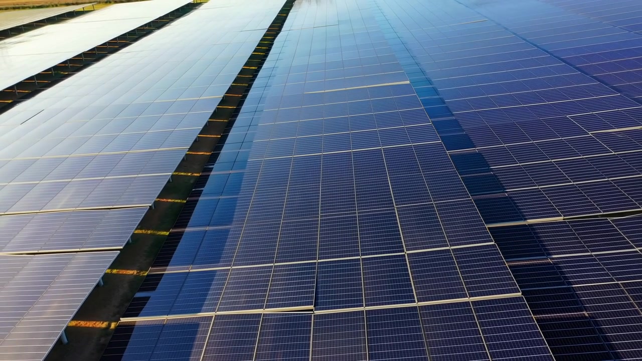 Una vista dall'alto si muove fluidamente su file parallele di pannelli fotovoltaici, con superfici blu riflettenti, linee a griglia geometriche, spazi in ombra e terreno illuminato dal sole che creano forti motivi lineari.
