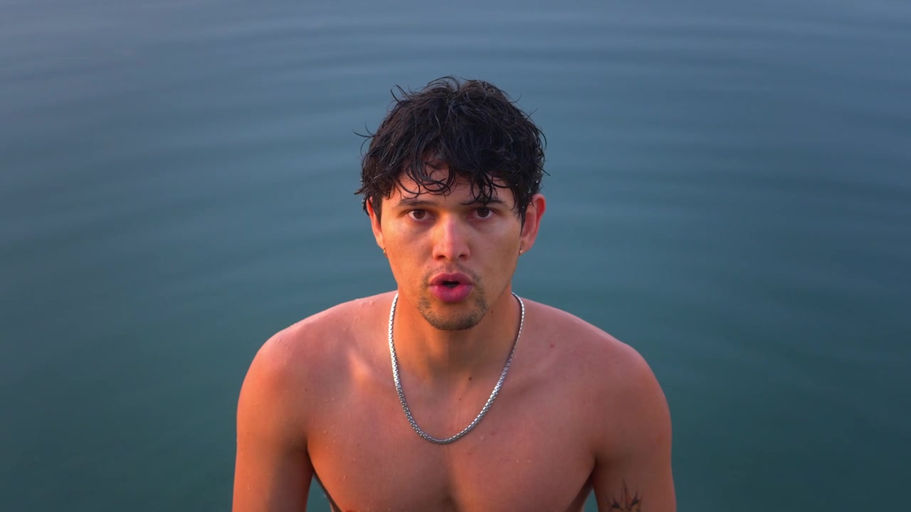 High-angle shot of a shirtless young Hispanic man standing waist-deep in calm lake water with subtle ripples reflecting the evening sky. Portrait shot, breathing technique.