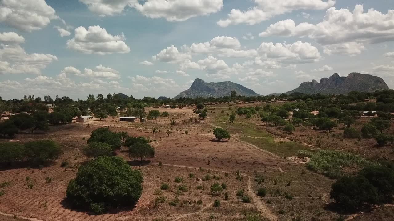 Rural landscape in northern Mozambique (Nampula region), with scattered trees, small settlements, and dramatic granite inselberg-style mountains rising in the distance under a cloud-filled sky