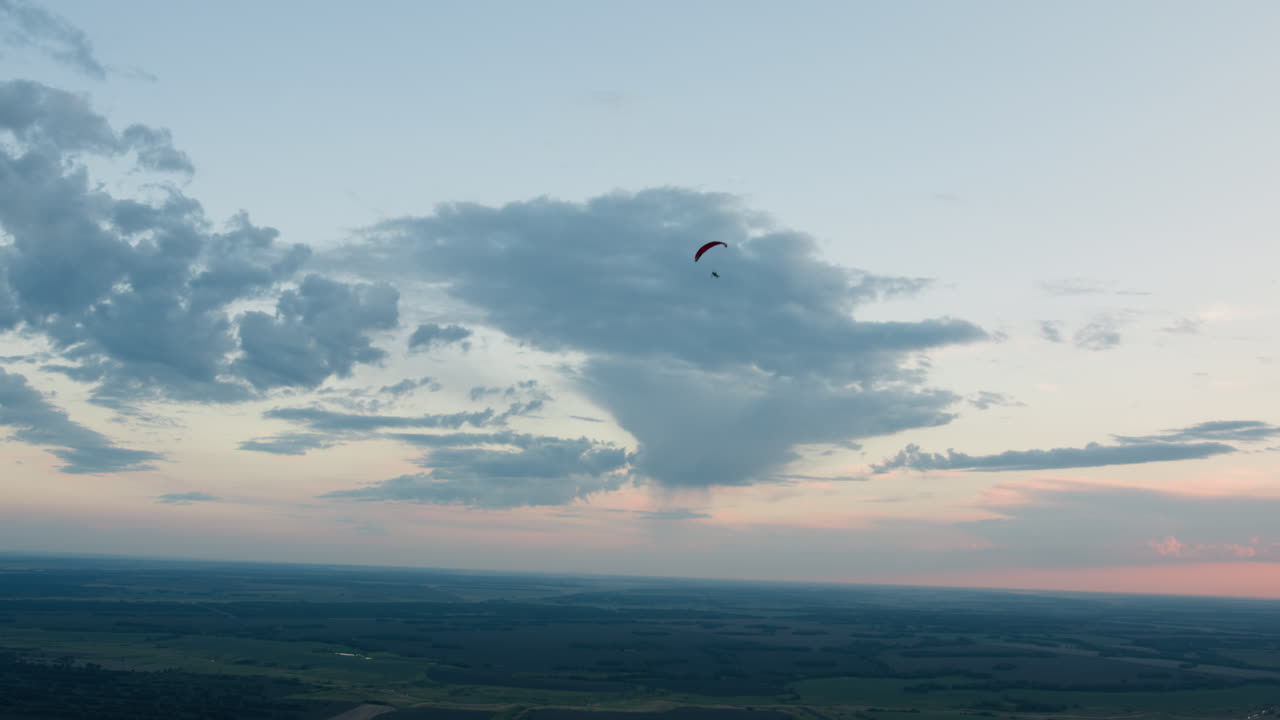 Distant silhouette of people flying powered paraglider under expansive sky with scattered clouds as they drift above farmland in gentle evening light conveying serene aerial motion