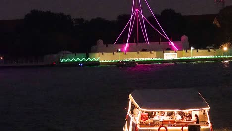 a vibrant boat passes a brightly lit structure with colorful lights reflecting on the water.