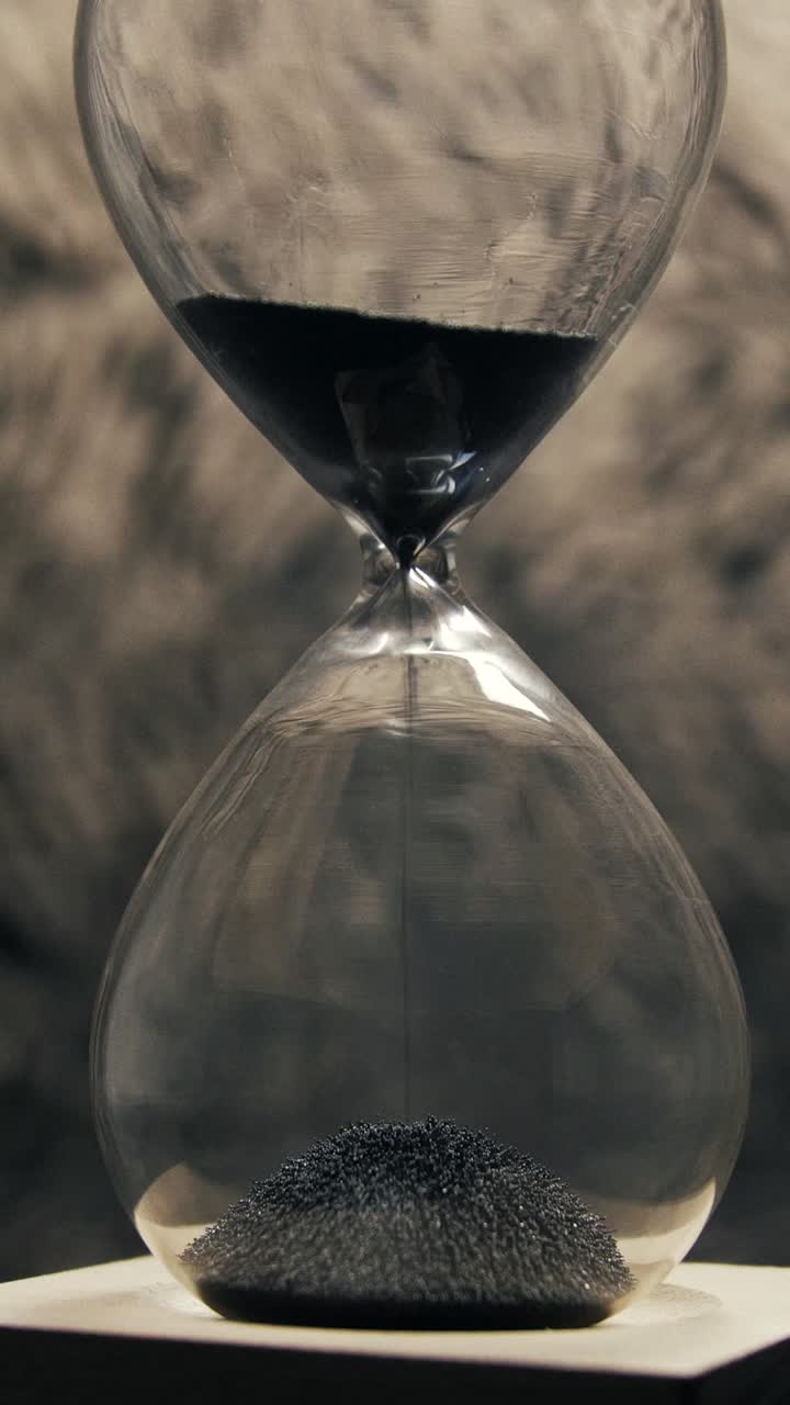 Clear glass hourglass with fine black sand steadily falling symbolising passing time and looming deadline