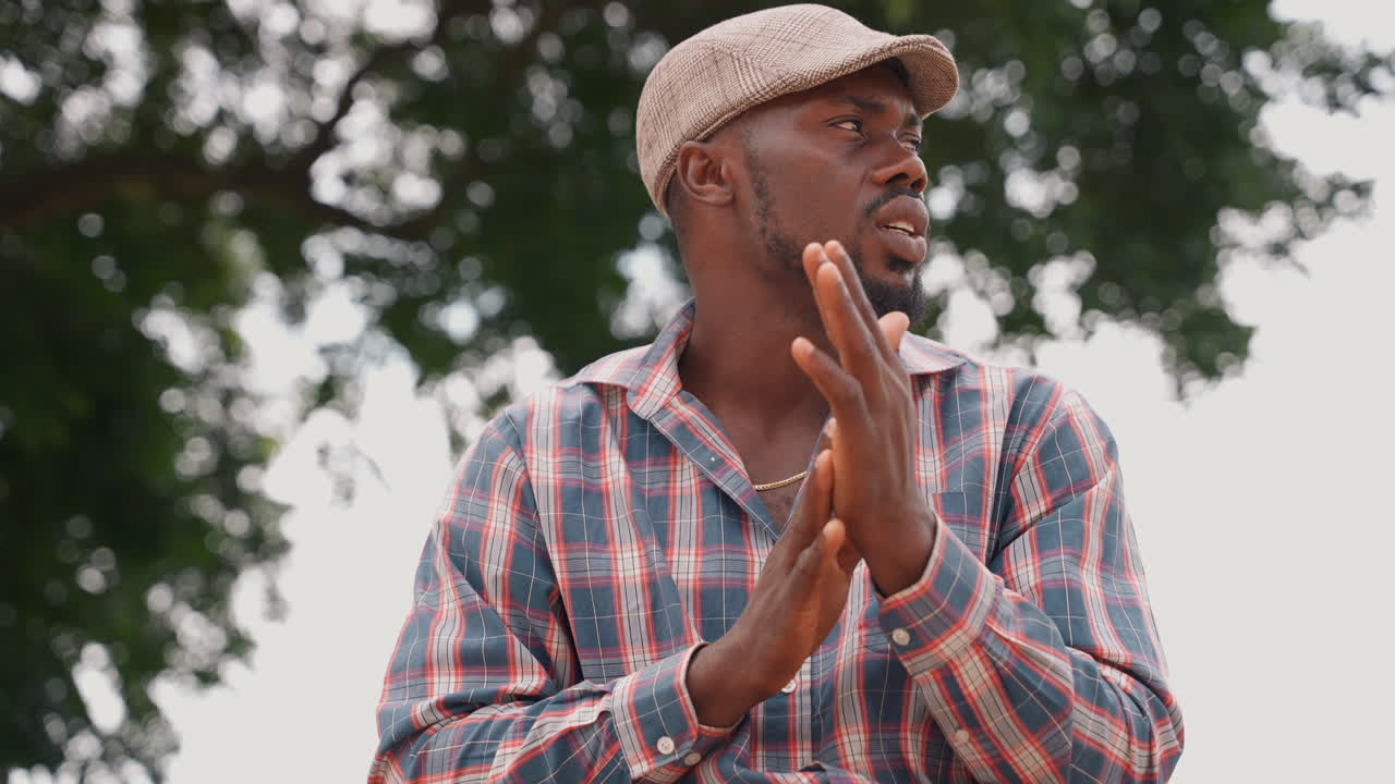 Outdoor man speaks with animated gestures, cap and patterned shirt under leafy canopy. Series of dynamic moments turning head, clapping hands, emphasizing points, briefly inspecting a small object