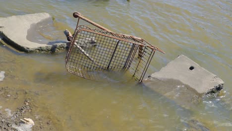 old and rusted shopping cart discarded in dirty river with chunks of broken concrete