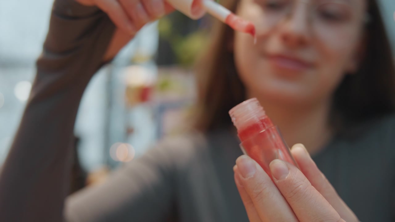 Manos sosteniendo un tubo de brillo labial rosa, quitando la tapa para mostrar el aplicador saturado, fondo de cafetería tipo invernadero con luces cálidas, primer plano espontáneo del producto y los dedos, luz natural, demostración de producto
