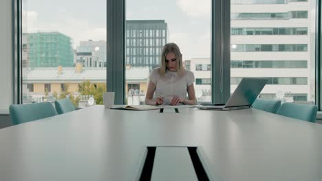 young businesswoman in conference room with papers. elegant blond woman sitting at long table in modern hall of office with laptop and writing on papers while working