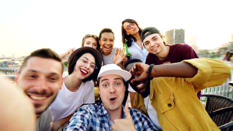 un joven guapo con gorra está mirando a la cámara y luego llama a sus amigos para que se tomen una selfie, la gente se está divirtiendo, haciendo caras y gestos divertidos y riendo.