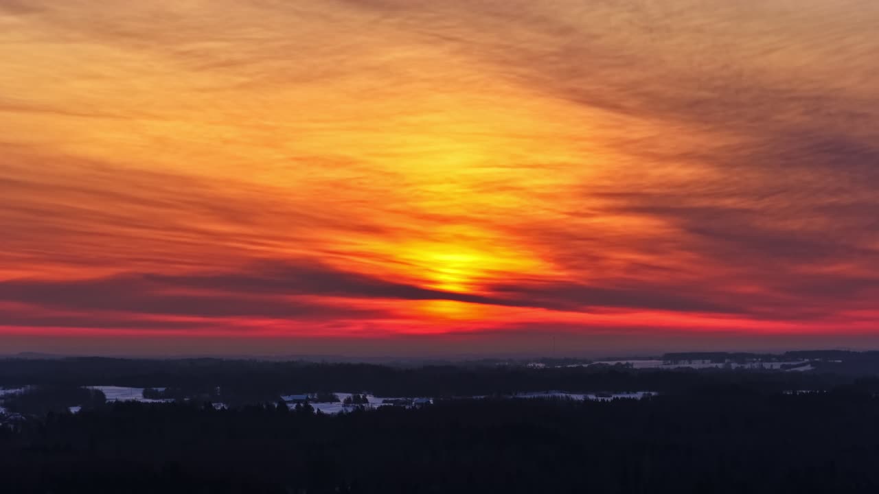 Dramatische Luftaufnahme des leuchtend orange-roten Sonnenuntergangs mit geschichteten Wolken über schneebedeckten Feldern und dunkler Waldsilhouette in Lettland