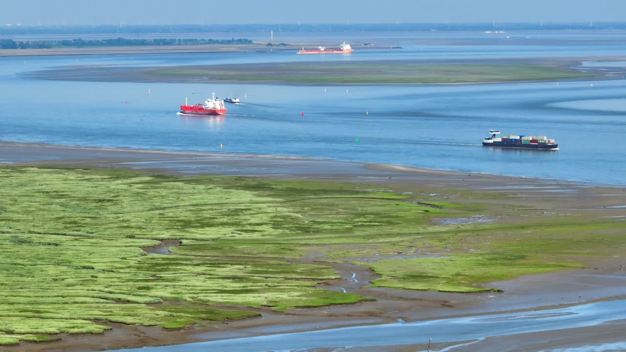 푸른 하늘 아래 녹색의 진흙 습지로 둘러싸인 넓은 강을 항해하는 화물선의 긴 공중 샷