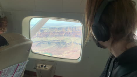 young caucasian woman sitting in window seat of airplane wearing face mask with scenic view of grand canyon colorful terrain, covid-19 pandemic, close up handheld