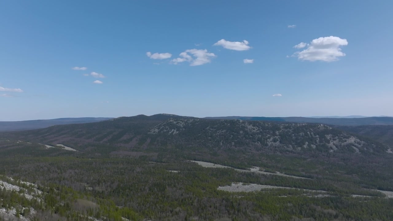 Drone footage of mountain ridge and vast taiga forest under blue sky with clouds in Taganay National Park near Zlatoust, Chelyabinsk Oblast, Russia