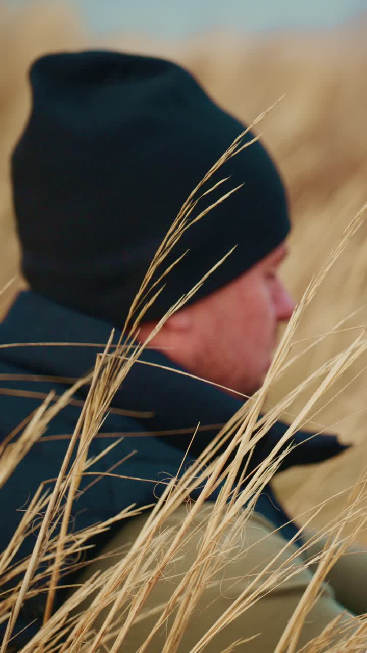 A reflective moment in nature: a man wearing a black beanie sits among tall golden grasses, showcasing a serene connection with the surrounding environment as he contemplates life in a peaceful outdoor setting
