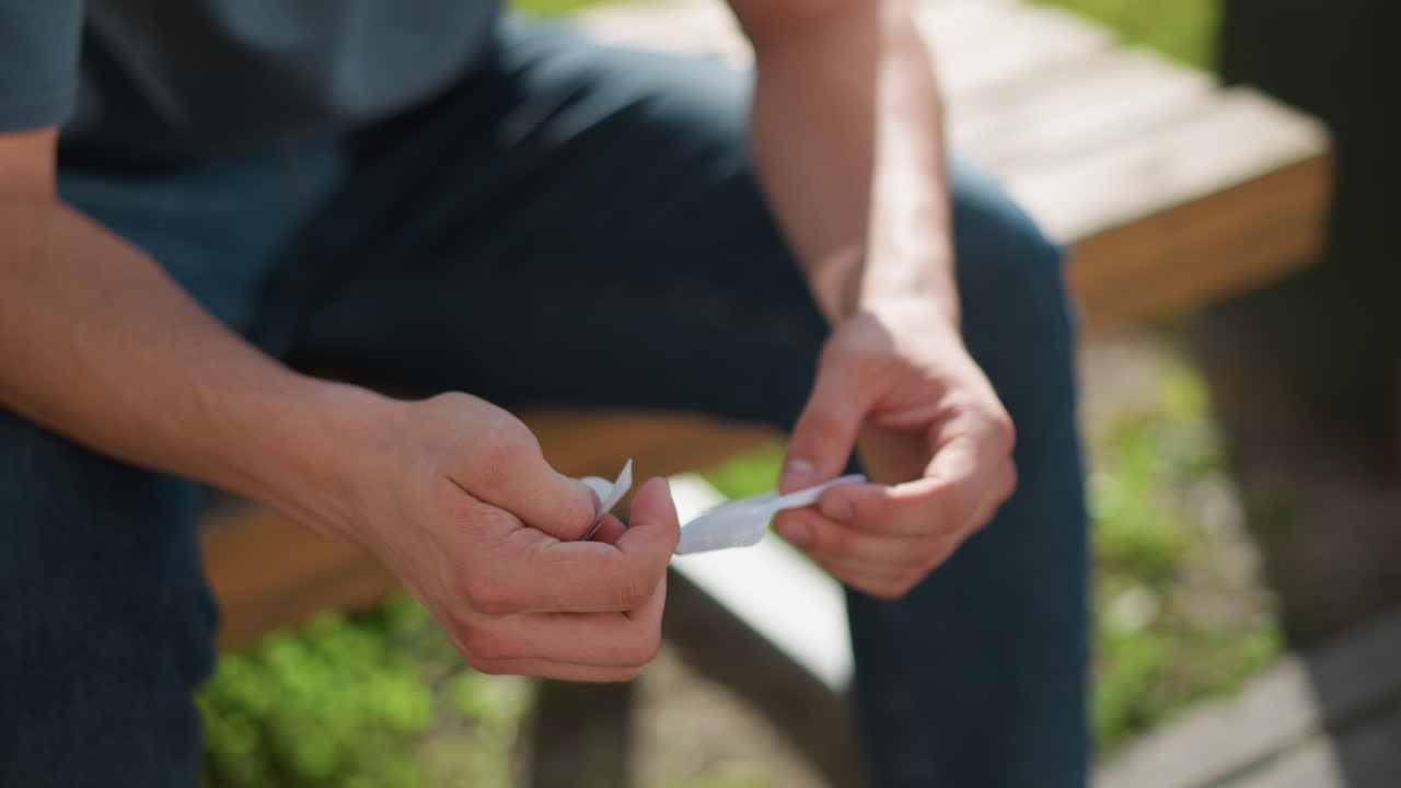 partial view of person seated placing nicotine patch on left arm while sitting on bench outdoors with soft greenery swaying gently in background