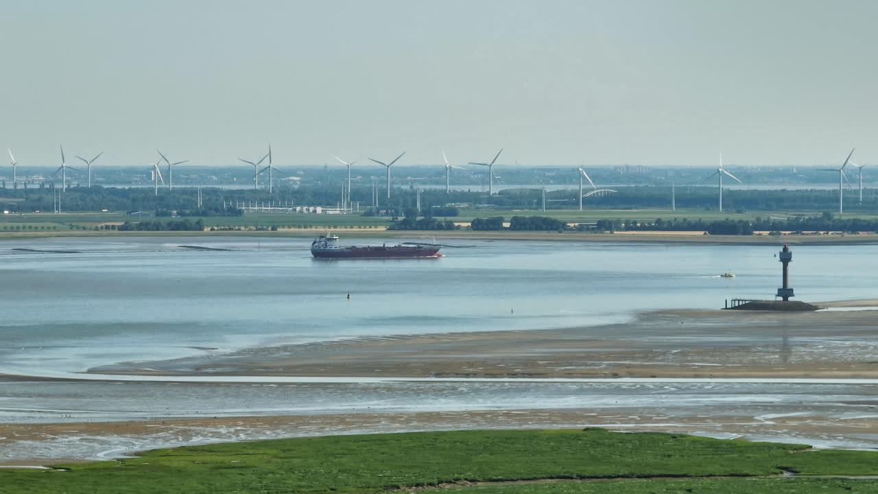 снимок с воздуха грузового судна, идущего по реке с заболоченной местностью на одной стороне и современной промышленностью с ветряными мельницами и заводами на другой стороне