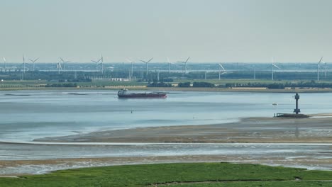 Aerial-shot-of-a-cargo-ship-navigating-a-river-with-wetlands-on-one-side-and-modern-industry-with-windmills-and-factories-on-the-other-side