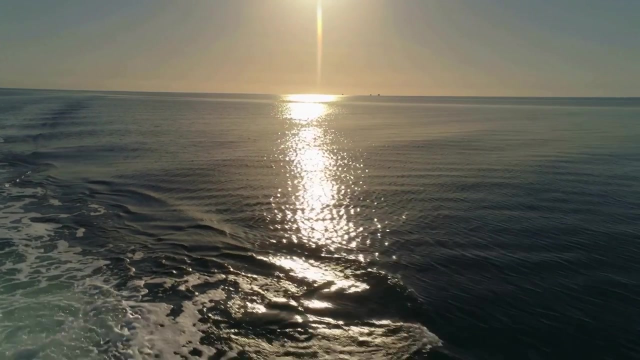 Lodret solrefleksion kastes over åbent hav, mens et fjerntliggende fartøj passerer og efterlader et skummende kølvand.