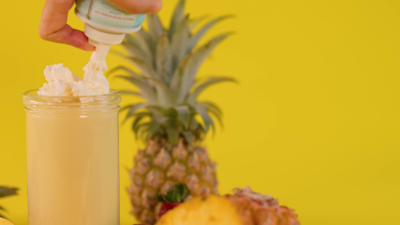 Una mano spruzza panna montata su un frullato all'ananas giallo in un barattolo di vetro