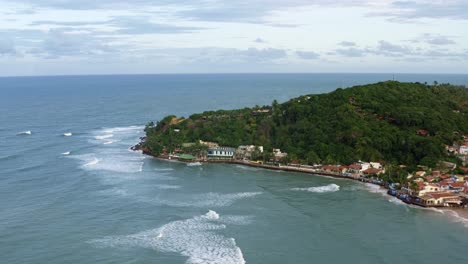 Rotating-aerial-drone-shot-of-the-beautiful-tropical-Pipa-beach-during-high-tide-with-small-waves-crashing-into-shore-surrounded-by-colorful-homes,-restaurants-and-green-exotic-foliage-on-a-summer-day