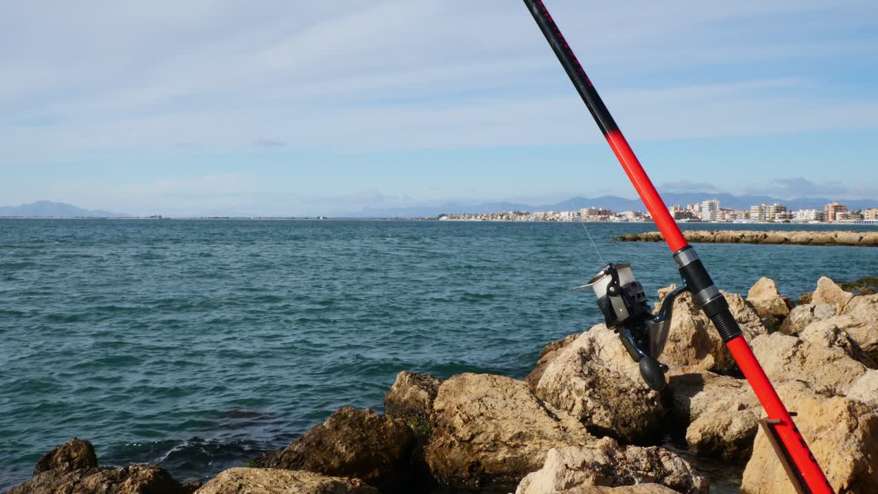Canna da pesca appoggiata sulle rocce con il villaggio sullo sfondo, Santa Pola, Spagna