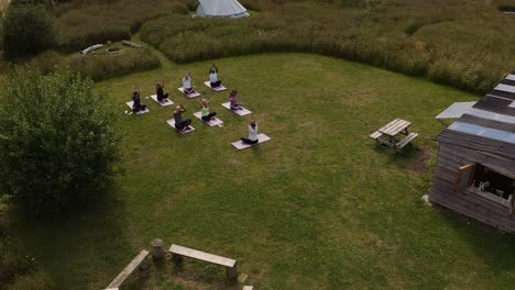 Drone-Shot-Of-Teacher-Leading-Group-Of-Mature-Men-And-Women-In-Class-At-Outdoor-Yoga-Retreat