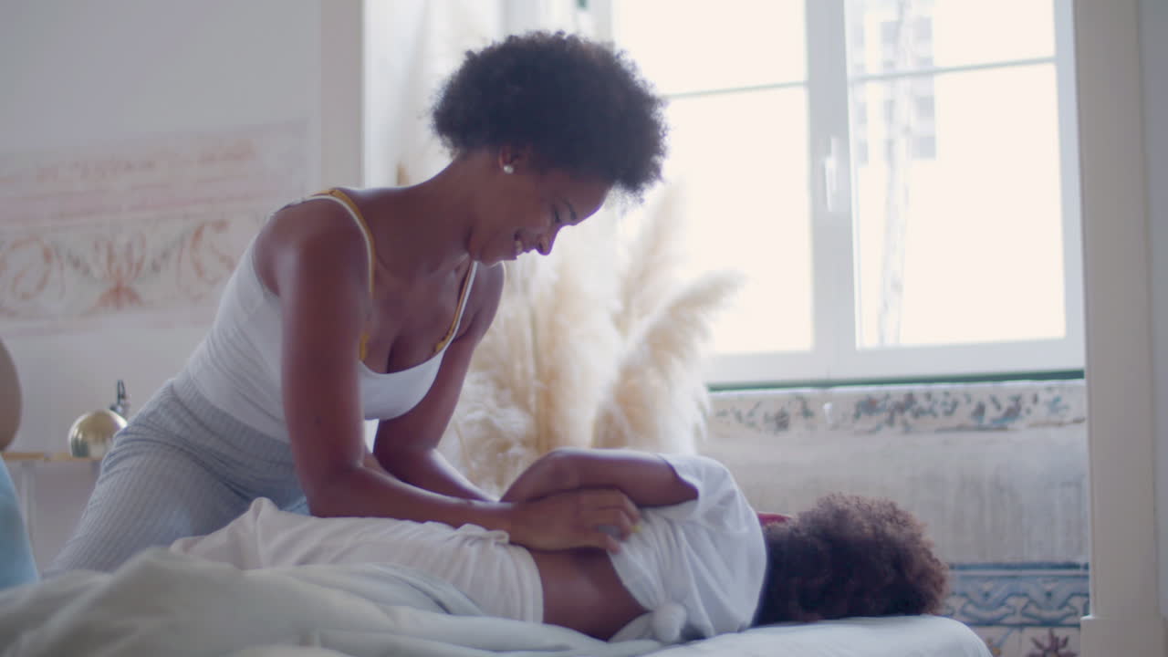 Mãe afro-americana fazendo cócegas na filhinha e se divertindo juntas na cama.
