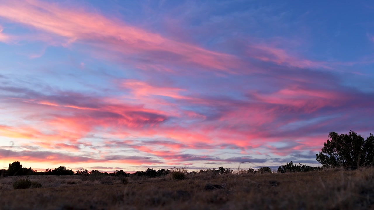 Timelapse 4K UHD che cattura un tramonto vivido. Nuvole rosa neon e blu brillanti si muovono rapidamente nel cielo, i loro colori svaniscono lentamente sopra un campo secco ed erboso nell'alto deserto