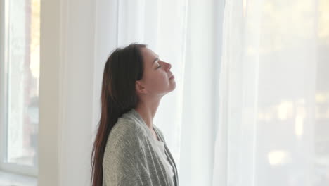 mujer con los ojos cerrados respirando profundamente mientras está de pie cerca de la ventana en casa por la mañana