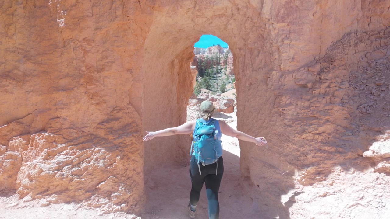 A woman with a backpack hikes through a sandstone tunnel in Bryce Canyon National Park. She spreads her arms as she walks through.