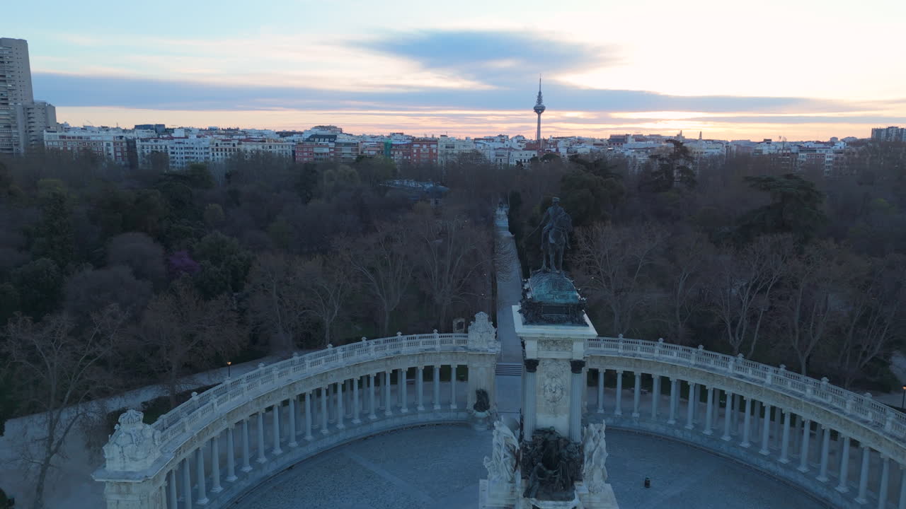 Breathtaking drone shot flying toward the statue at Retiro Park's pond during sunrise. The skyline, including the iconic Torrespaña tower, is visible in the background, with warm morning light