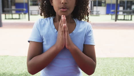 biracial girl with curly hair is in a praying pose outdoors at school