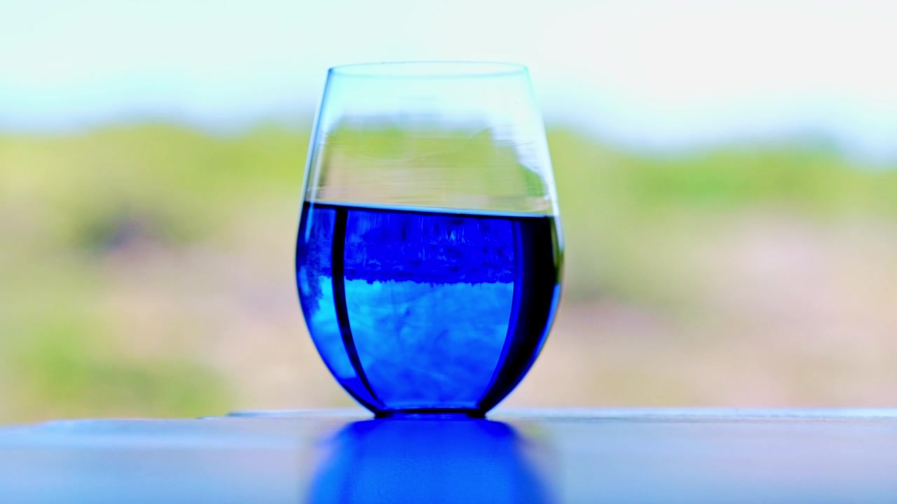 Un vaso con Azul de Metileno arremolinándose diluido en agua sobre una mesa frente a una ventana.