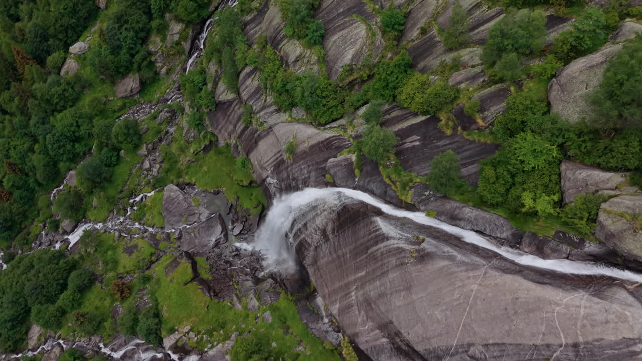 Drone follows a waterfall cascading down steep rocks in the Alps, then orbits around it, revealing the flow in a full top-down view