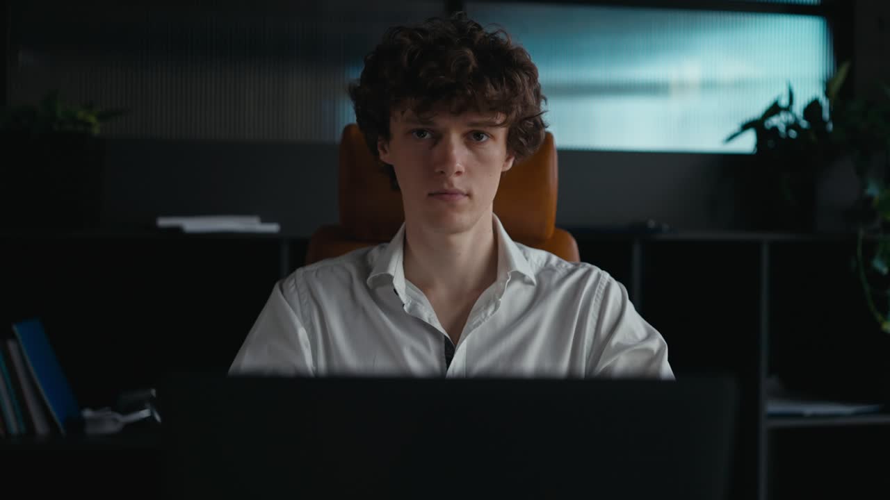 Portrait of a confident guy with curly hair in a white shirt who works at a gray laptop in the office and looks at the camera without a smile on his face