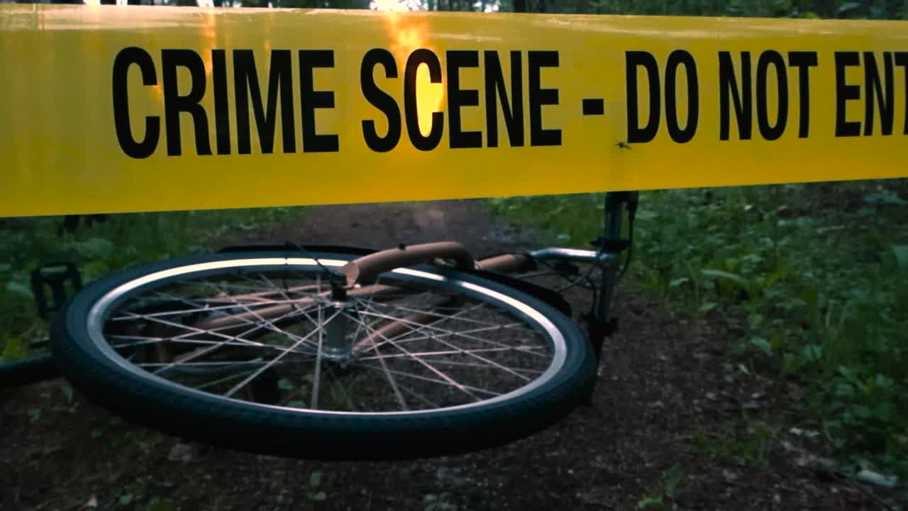 Low angle of fallen bicycle behind the crime scene tape, accident scene. Bike wheel spinning lying on the ground on summer evening forest trail. Incident site on wooded area road. True crime setting